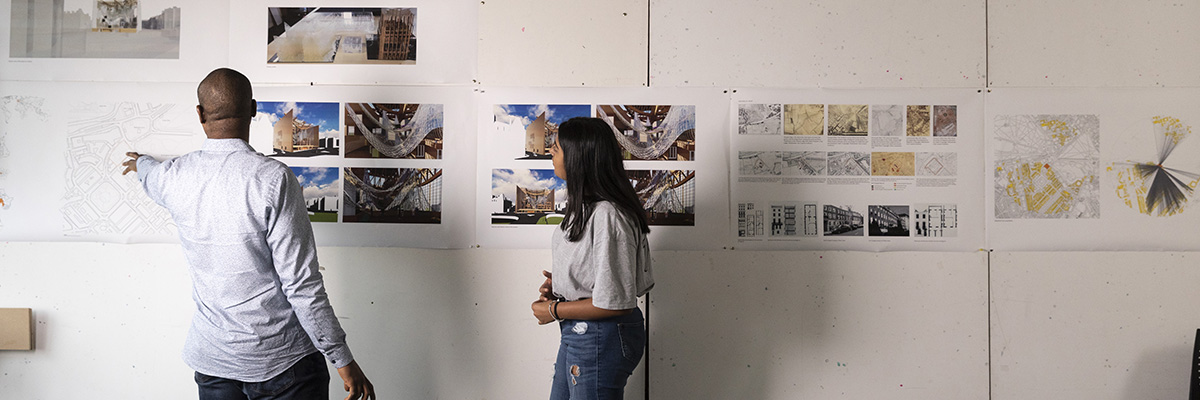A lecturer points his student toward a blueprint pinned on a wall full of structural images